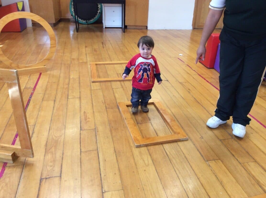 Niño pequeño de pie sobre una tabla de madera del circuito, con su maestra a un costado