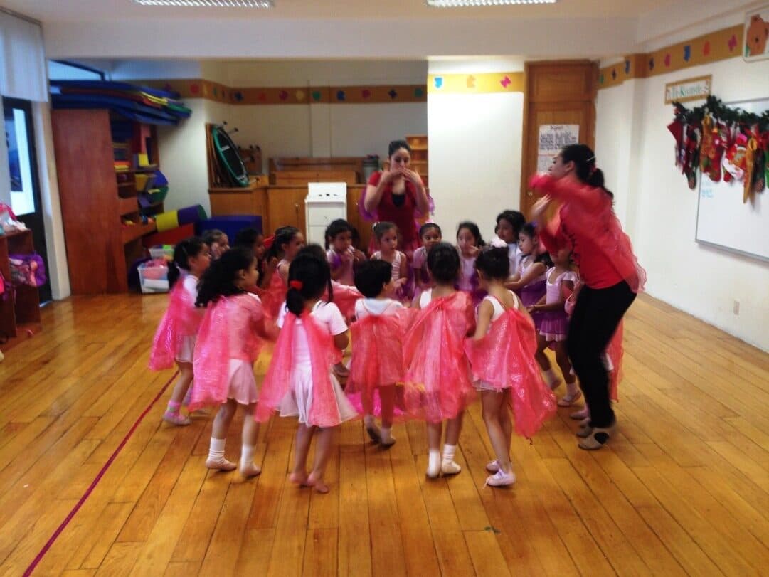 Alumnas con tutús rosas bailando en círculo alrededor de su maestra de ballet