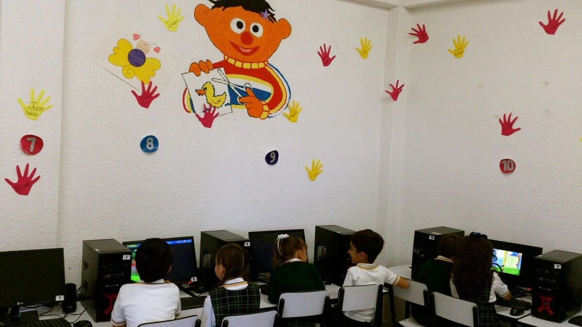 Alumnos trabajando en las computadoras del aula de cómputo, con un mural infantil en la pared